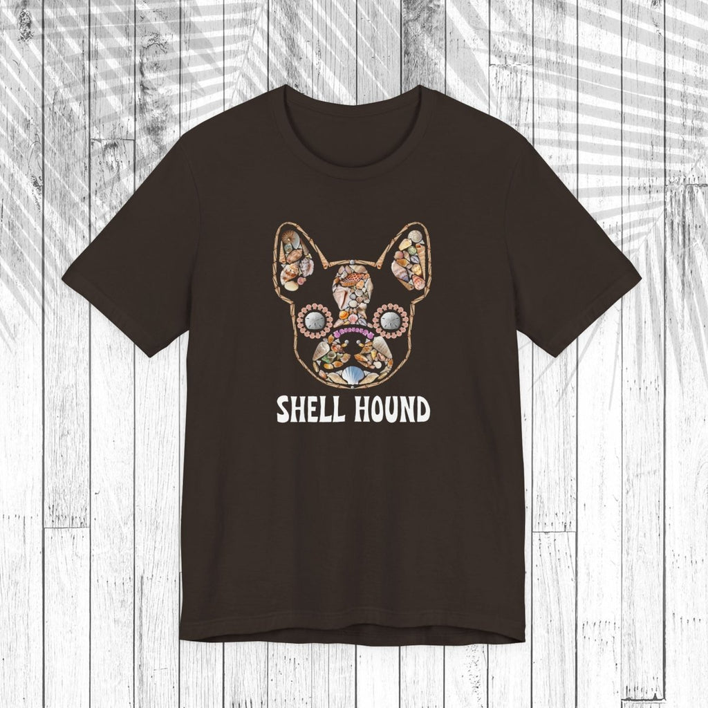 A dark brown short-sleeve t-shirt with a graphic of a French Bulldog decorated with a colorful floral pattern and the text 'SHELL HOUND' printed below in white, displayed on a white wood plank background.
