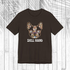 A dark brown short-sleeve t-shirt with a graphic of a French Bulldog decorated with a colorful floral pattern and the text 'SHELL HOUND' printed below in white, displayed on a white wood plank background.