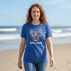 A woman with red hair stands on a sandy beach wearing a heather blue graphic t-shirt featuring a French Bulldog design with 'SHELL HOUND' printed below it.