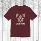 A dark brown short-sleeve t-shirt with a graphic of a French Bulldog decorated with a colorful floral pattern and the text 'SHELL HOUND' printed below in white, displayed on a white wood plank background.