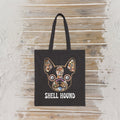 A black canvas tote bag with a colorful mosaic frenchton bulldog face graphic using seashells  and the text 'SHELL HOUND' printed below it, displayed against a rustic whitewashed wooden wall.
