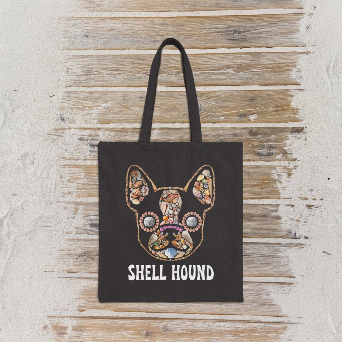 A black canvas tote bag with a colorful mosaic frenchton bulldog face graphic using seashells  and the text 'SHELL HOUND' printed below it, displayed against a rustic whitewashed wooden wall.