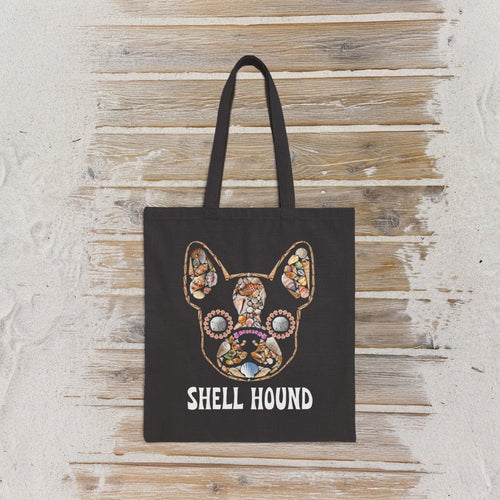 A black canvas tote bag with a colorful mosaic frenchton bulldog face graphic using seashells  and the text 'SHELL HOUND' printed below it, displayed against a rustic whitewashed wooden wall.