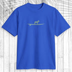 A royal blue short-sleeve crewneck t-shirt featuring a light sage green graphic of a dog standing on a surfboard across the chest, displayed flat against a white wood plank background.