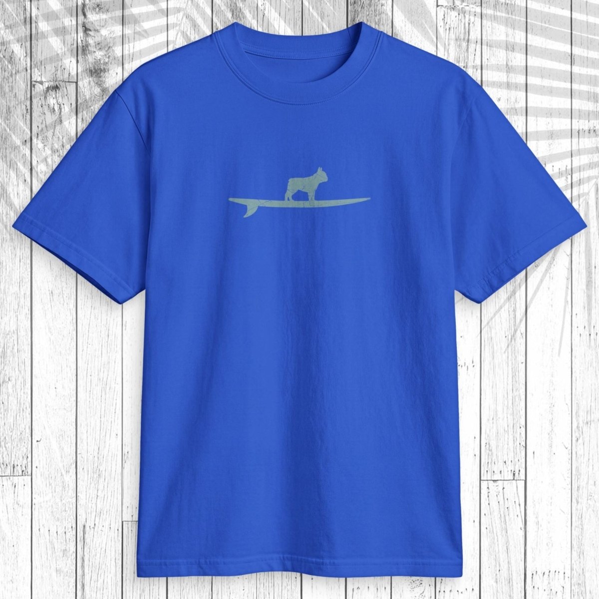 A royal blue short-sleeve crewneck t-shirt featuring a light sage green graphic of a dog standing on a surfboard across the chest, displayed flat against a white wood plank background.