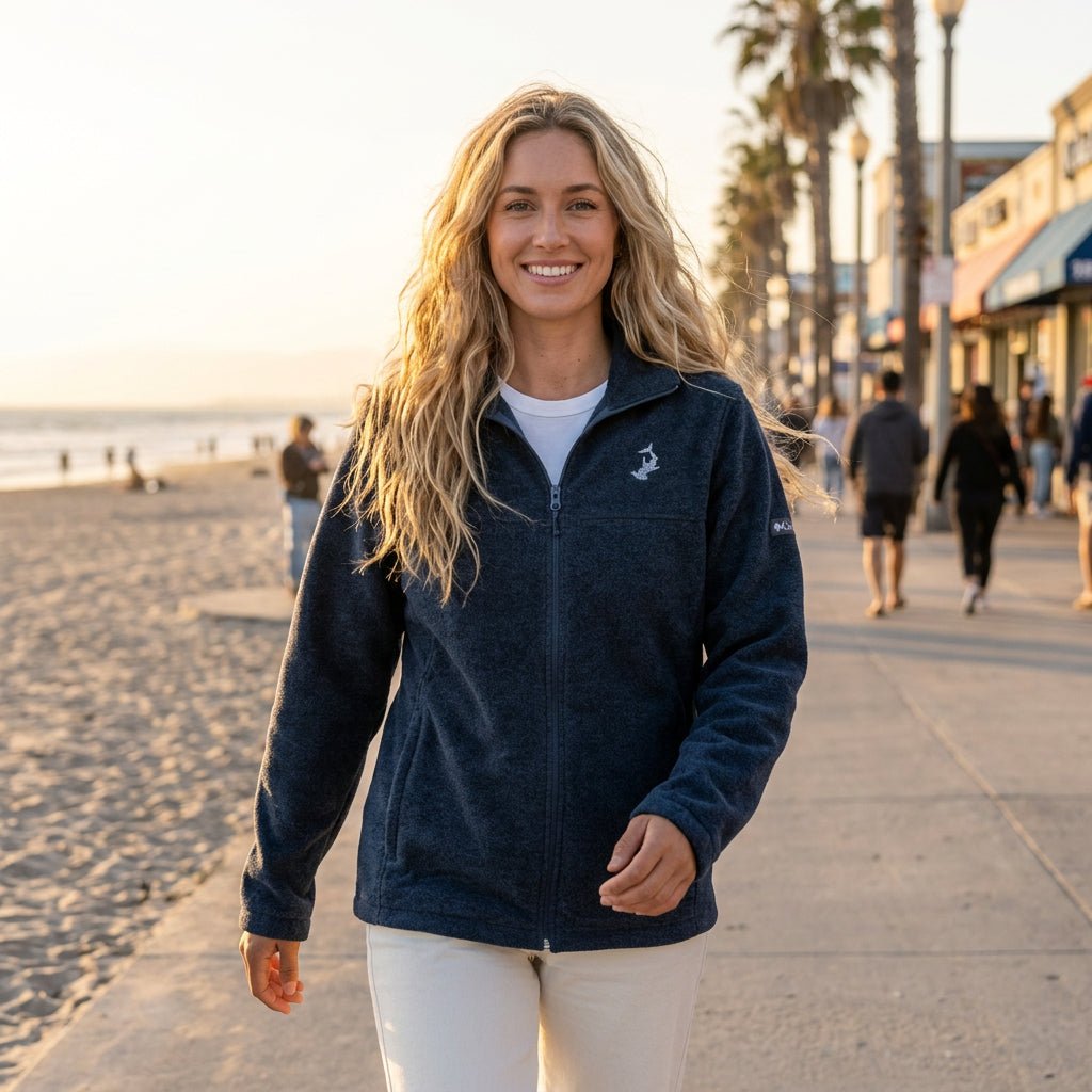 A woman stands on a sandy beach wearing a dark navy heathered zip-up fleece jacket with a small white embroidered logo on the left chest, paired with off-white pants.