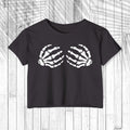 A dark charcoal gray short-sleeve crop top t-shirt featuring a white graphic of two skeletal hands on the chest, displayed flat against a white wood plank background.