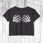 A dark charcoal gray short-sleeve crop top t-shirt featuring a white graphic of two skeletal hands on the chest, displayed flat against a white wood plank background.