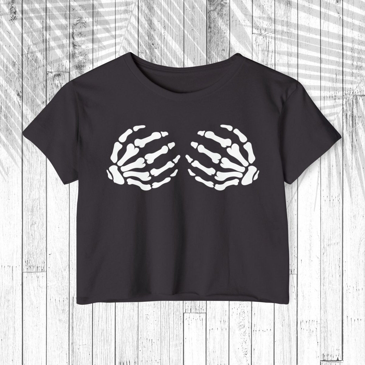 A dark charcoal gray short-sleeve crop top t-shirt featuring a white graphic of two skeletal hands on the chest, displayed flat against a white wood plank background.
