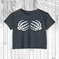 A dark charcoal gray short-sleeve crop top t-shirt featuring a white graphic of two skeletal hands on the chest, displayed flat against a white wood plank background.