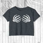 A dark charcoal gray short-sleeve crop top t-shirt featuring a white graphic of two skeletal hands on the chest, displayed flat against a white wood plank background.