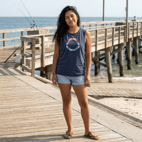 A woman stands on a wooden pier by the ocean wearing a navy blue sleeveless tank top with a colorful Shark Jaw graphic on the front, paired with light denim shorts and brown sandals as she is ready to go fishing.