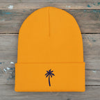 A bright gold ribbed knit beanie hat with a folded cuff and a small dark blue palm tree embroidered on the front, shown against a plain rustic wood background.