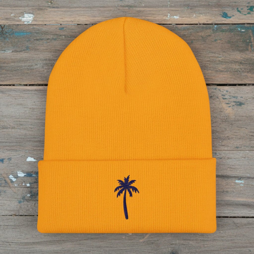 A bright gold ribbed knit beanie hat with a folded cuff and a small dark blue palm tree embroidered on the front, shown against a plain rustic wood background.