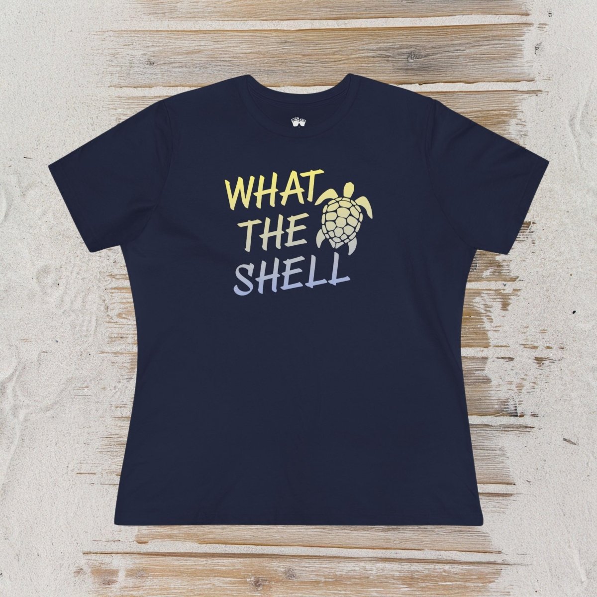 A navy blue short-sleeve t-shirt with a graphic print reading 'WHAT THE SHELL' in yellow and light blue, accompanied by a sea turtle illustration, laid flat on a light wood plank surface.