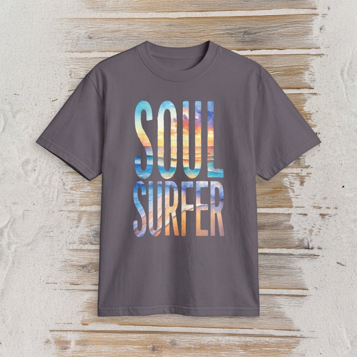 A dark gray crewneck t-shirt featuring a colorful graphic print that reads 'SOUL SURFER' in a watercolor sunset gradient, displayed flat against a rustic light wood background.