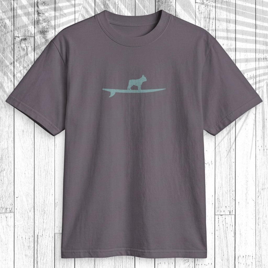 A dark gray crewneck t-shirt featuring a light teal graphic of a dog standing on a surfboard across the chest, displayed flat against a white wood plank background with a palm leaf shadow overlay.
