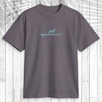 A dark gray crewneck t-shirt featuring a light teal graphic of a dog standing on a surfboard across the chest, displayed flat against a white wood plank background with a palm leaf shadow overlay.