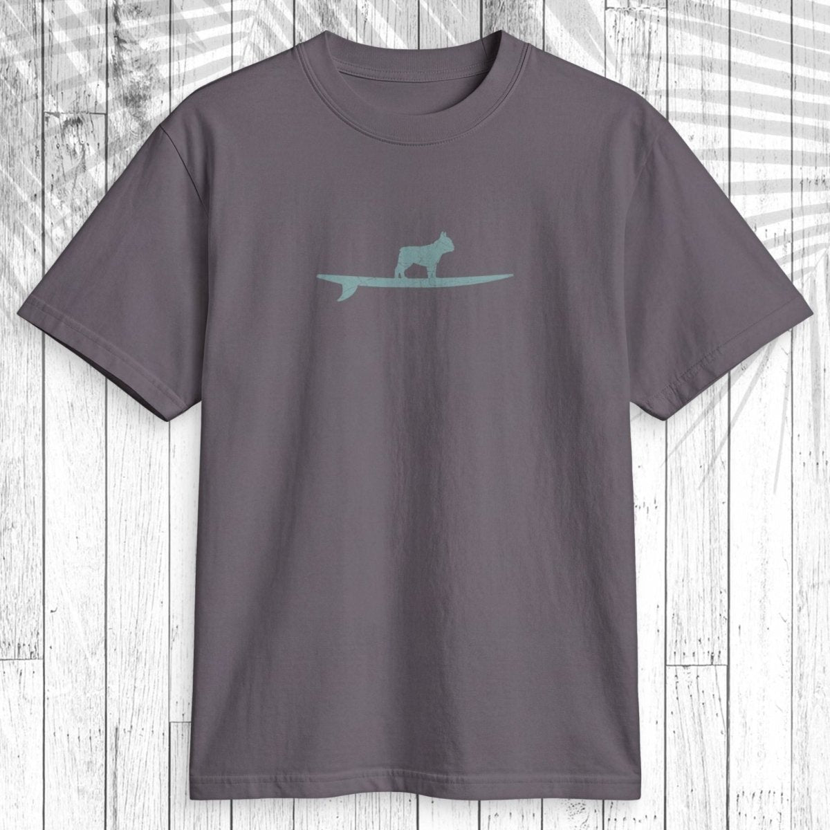 A dark gray crewneck t-shirt featuring a light teal graphic of a dog standing on a surfboard across the chest, displayed flat against a white wood plank background with a palm leaf shadow overlay.