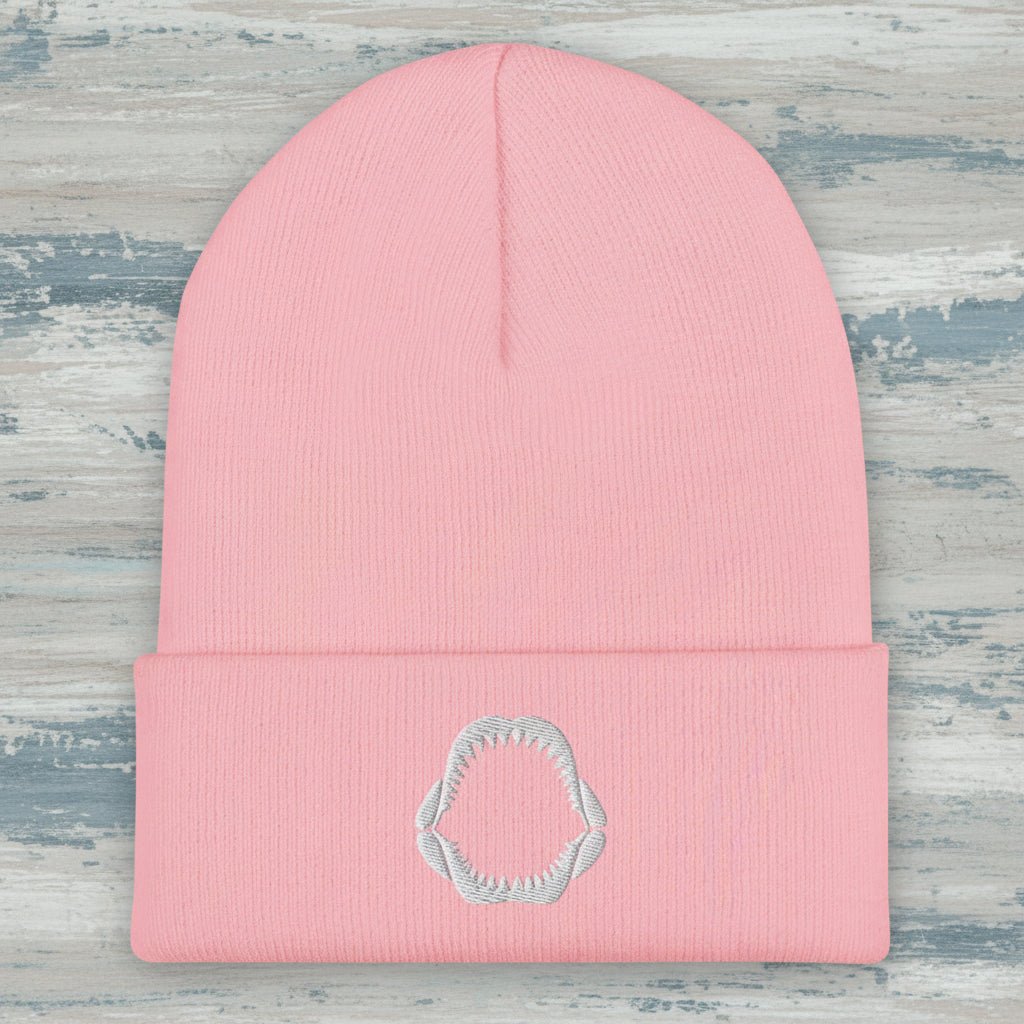 A light pink ribbed knit beanie hat with a folded cuff and a small white embroidered shark jaw embroidery on the front, shown against a rustic wood background