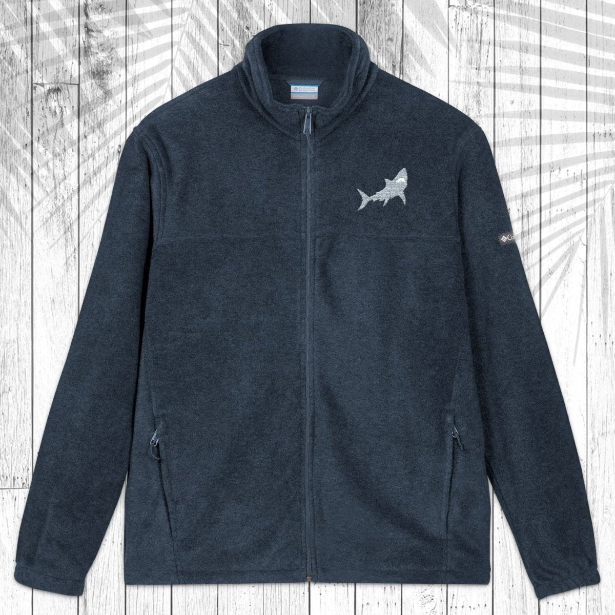 A dark gray fleece zip-up jacket with a stand collar, shown flat against a white wood plank background with a palm leaf pattern overlay. The jacket features a Great White Shark embroidered on the left chest and a small white logo on the right sleeve.