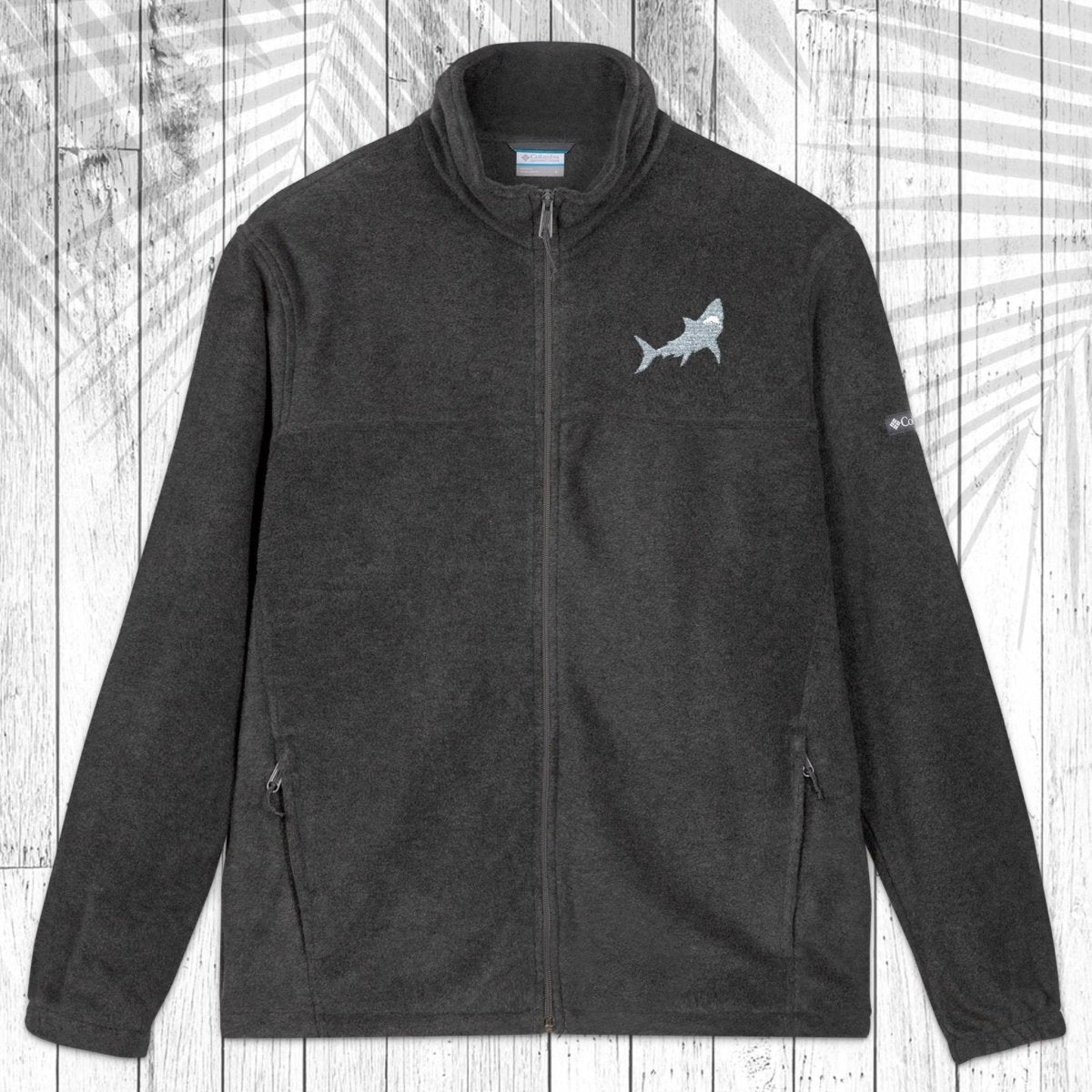 A dark gray fleece zip-up jacket with a stand collar, shown flat against a white wood plank background with a palm leaf pattern overlay. The jacket features a small white circular logo on the left chest and a small white logo on the right sleeve.