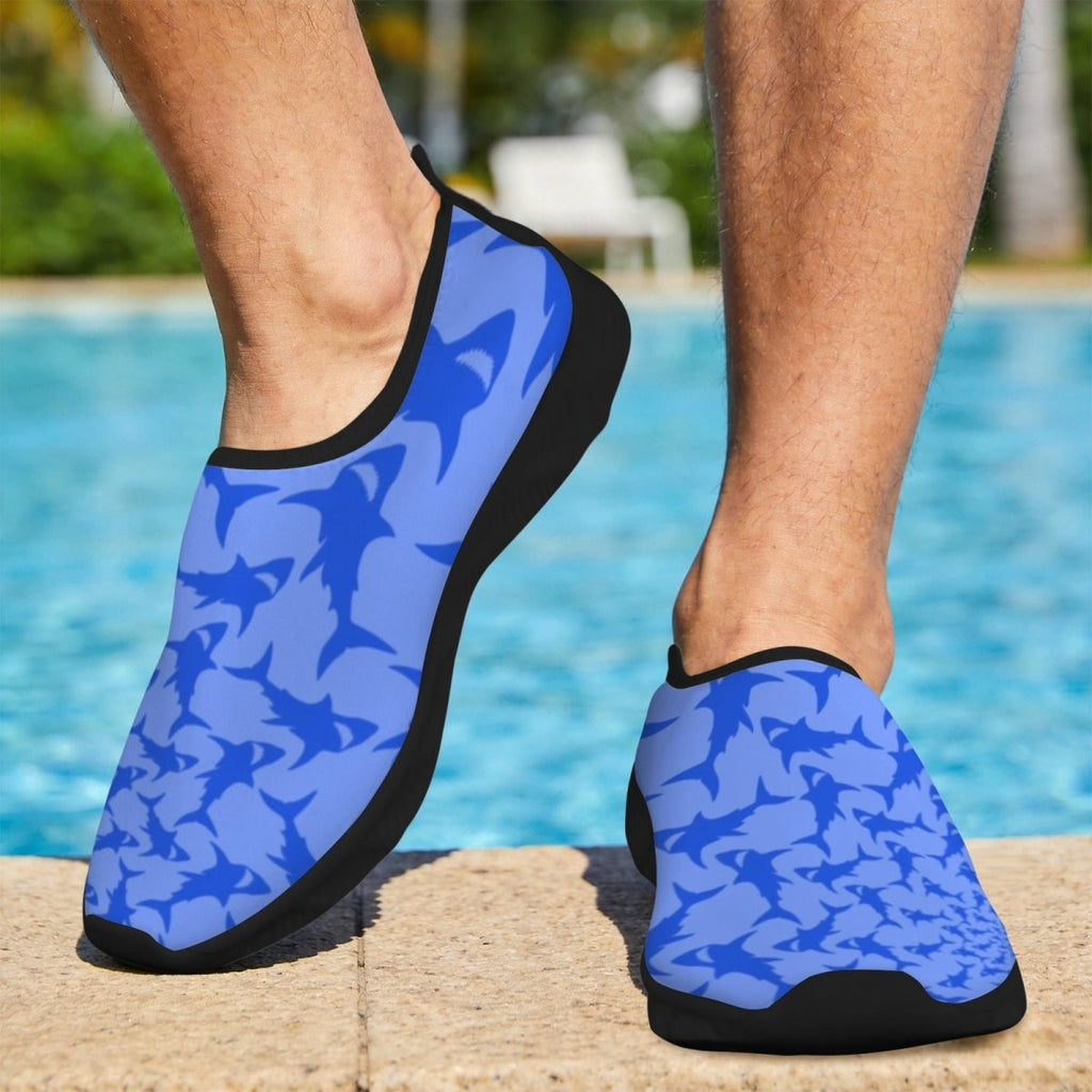 A person wearing blue and light blue shark-patterned water shoes with black soles, standing on a pool deck with a swimming pool in the background.