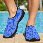 A person wearing blue and light blue shark-patterned water shoes with black soles, standing on a pool deck with a swimming pool in the background.