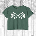 A dark green heathered short-sleeve crop top featuring a white graphic of two skeletal hands reaching toward each other, displayed flat against a white wood plank background.