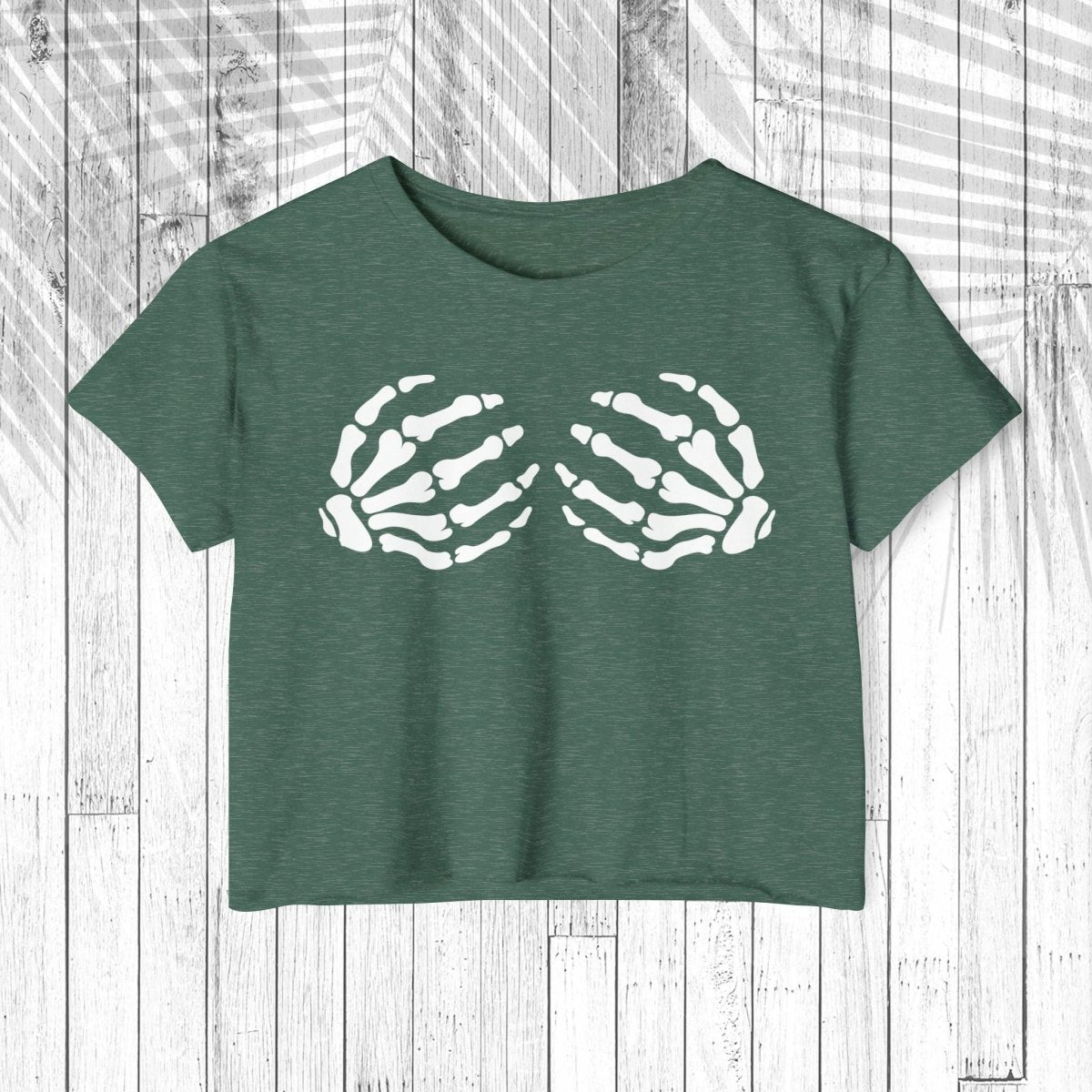 A dark green heathered short-sleeve crop top featuring a white graphic of two skeletal hands reaching toward each other, displayed flat against a white wood plank background.