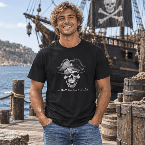 A smiling man wearing a black graphic t-shirt stands on a wooden dock with a pirate ship and sea in the background. The t-shirt features a grayscale graphic of a skull wearing a pirate hat with the text 'You Should Know How To Use A Sword'.