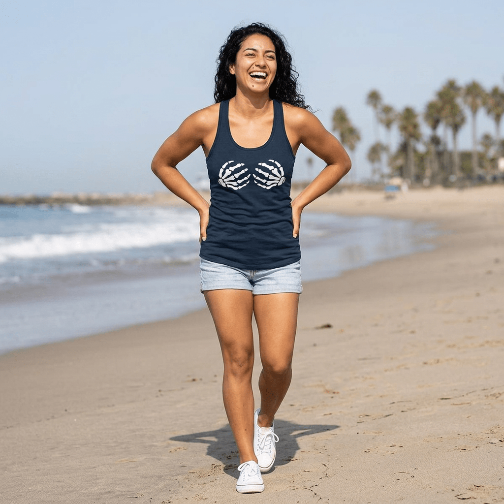 A woman smiling and walking on a sandy beach wearing a navy blue tank top with a white graphic design, light denim shorts, and white sneakers.