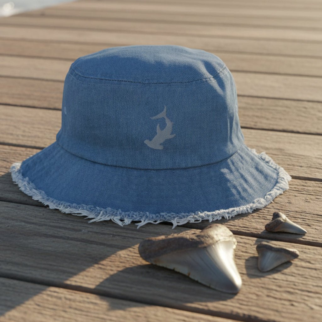 Blue denim bucket hat with Hammerhead shark embroidery sitting on a beachside boardwalk with shark teeth laying around