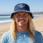Man wearing a blue bucket hat with a shaka bones logo, smiling against a white background 