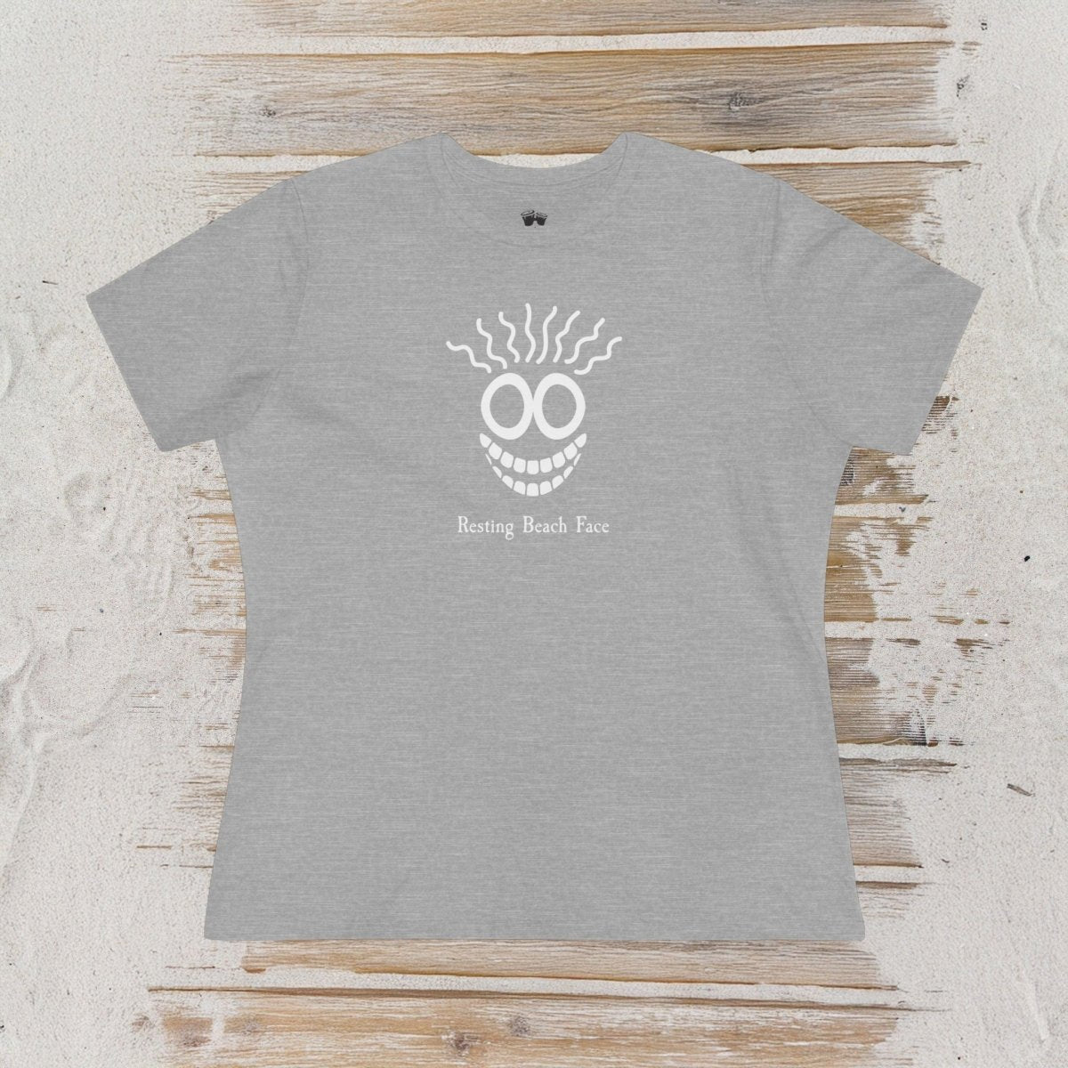 A heather gray short-sleeve t-shirt with a white graphic of a smiling face with teeth and the text 'Betting Beach Face' printed on the chest, laid flat on a rustic light wood background.