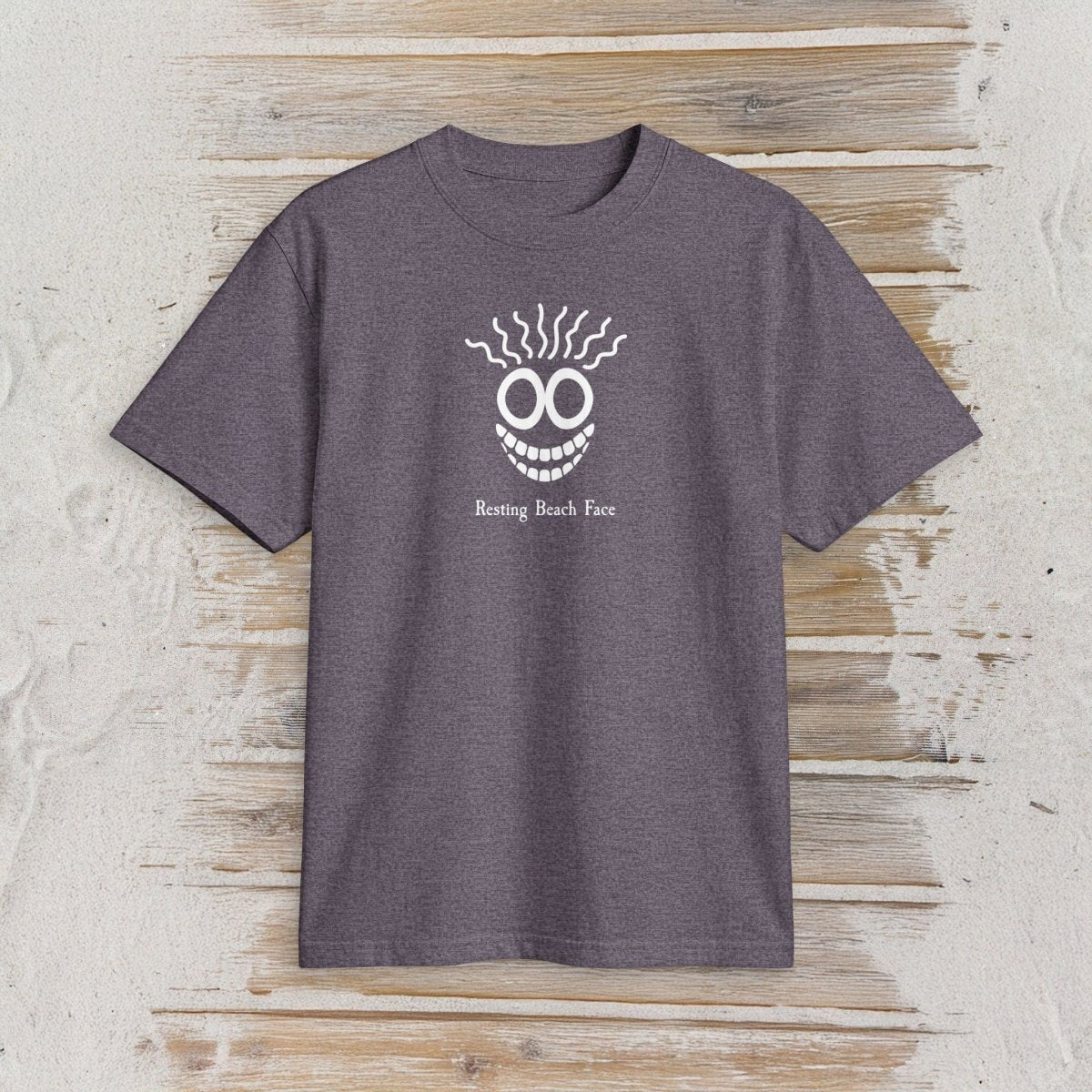 A heather dark purple short-sleeve t-shirt with a white graphic of a smiling face with wavy hair and the text 'Resting Beach Face' printed on the chest, displayed flat on a rustic light wood background.