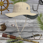 Beige sun hat with embroidered skull and crossbones design on a wooden background with pirate-themed items.