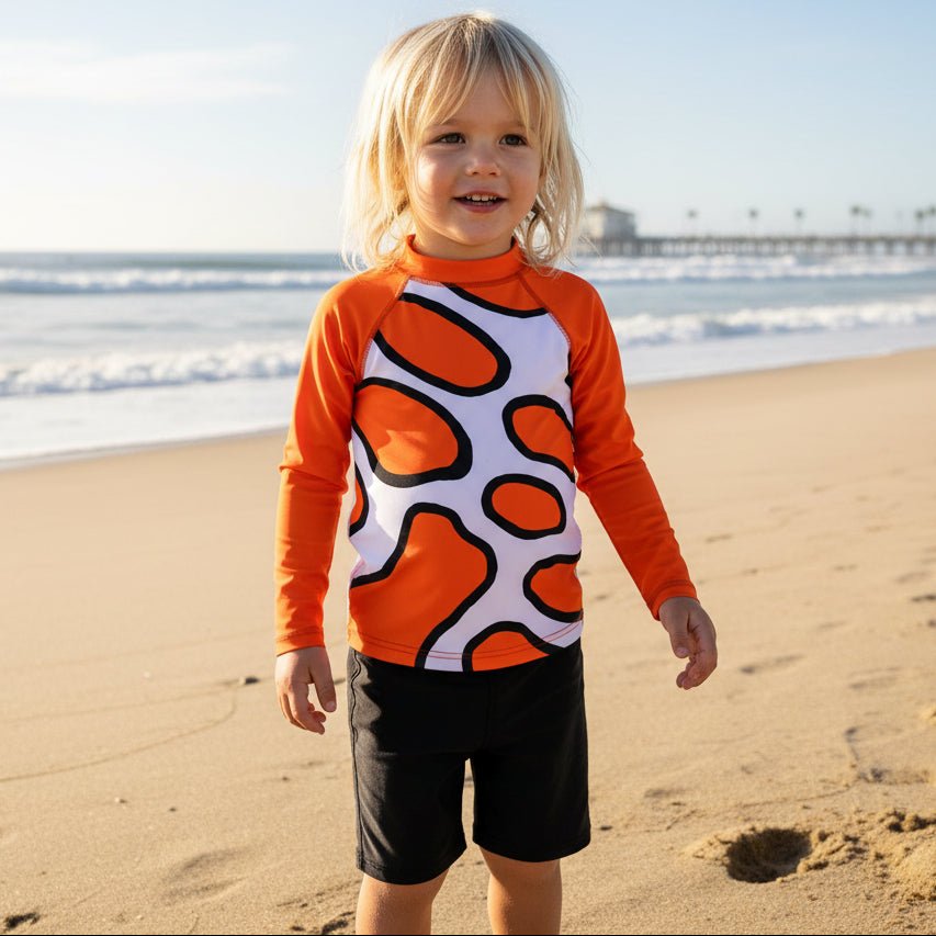 A young child standing on hermosa beach california, wearing a long-sleeve swim shirt, with an orange, white, and black abstract pattern, paired with black swim shorts.