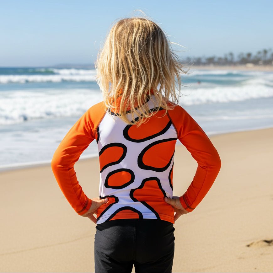 A child standing on Hermosa Beach with their back to the camera, wearing a long-sleeve rash guard with a white body, orange sleeves, and an orange and black abstract pattern.