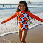 A young girl smiling on Hermosa beach with her arms outstretched, wearing a long-sleeve swim rashie featuring a white base with bold orange abstract Clown Fish spots outlined in black, and bright orange sleeves.
