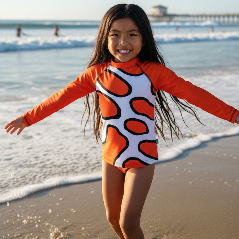 A young girl smiling on Hermosa beach with her arms outstretched, wearing a long-sleeve swim rashie featuring a white base with bold orange abstract Clown Fish spots outlined in black, and bright orange sleeves.