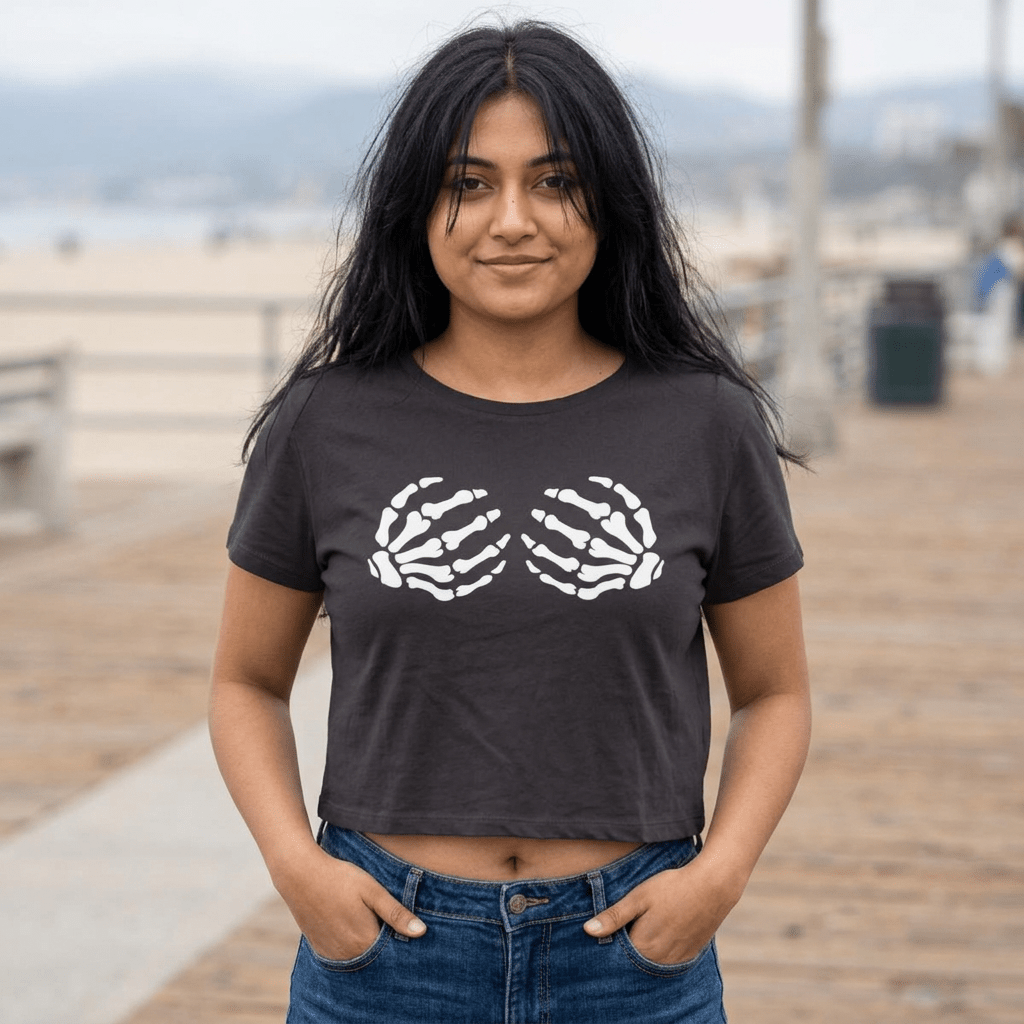 A woman wearing a dark charcoal gray crop top t-shirt with a white graphic of two skeletal hands, standing on a wooden boardwalk with a blurred beach background.
