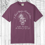 A short-sleeve crewneck t-shirt in a dusty plum purple color, featuring a white graphic of a skull wearing a pirate hat with the text 'I Tried Being Normal Once' above and 'It Was The Worst 2 Minutes of My Life' below. The t-shirt is displayed flat against a white wood plank background.