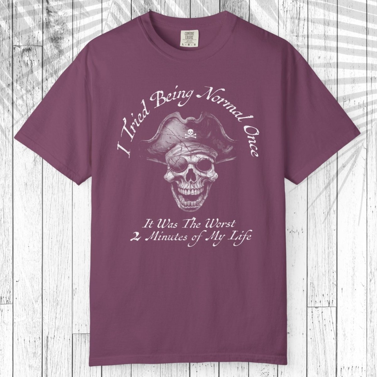 A short-sleeve crewneck t-shirt in a dusty plum purple color, featuring a white graphic of a skull wearing a pirate hat with the text 'I Tried Being Normal Once' above and 'It Was The Worst 2 Minutes of My Life' below. The t-shirt is displayed flat against a white wood plank background.