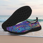 A pair of slip-on water shoes with a vibrant multicolor psychedelic pattern on the upper and black soles, displayed on a sandy beach with the ocean in the background.