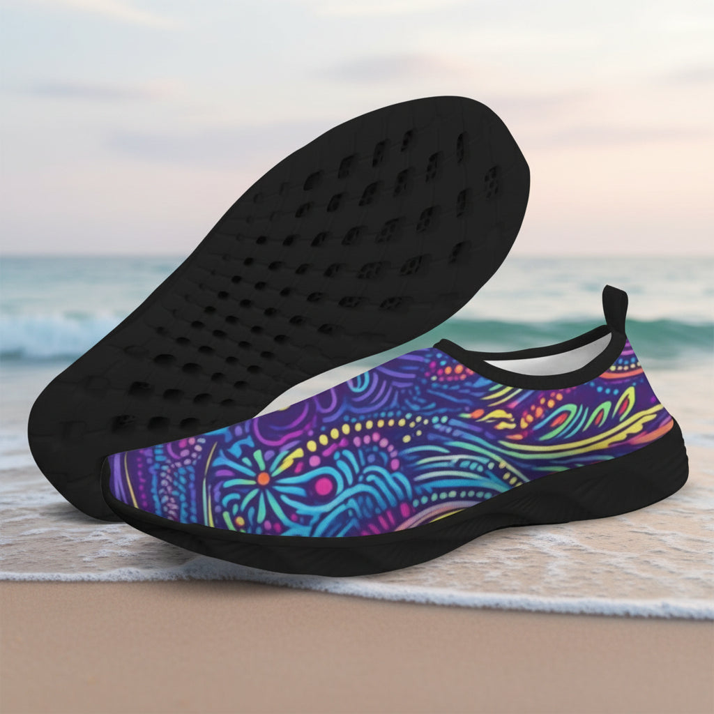A pair of slip-on water shoes with a vibrant multicolor psychedelic pattern on the upper and black soles, displayed on a sandy beach with the ocean in the background.