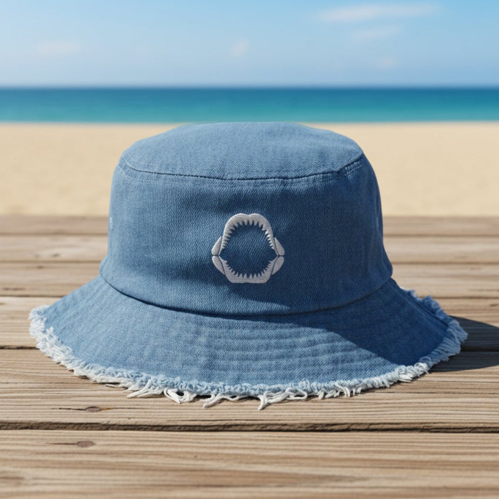 Blue denim bucket hat with an embroidered shark jaw sitting on a beachside boardwalk