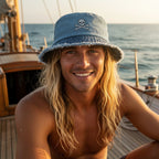 Dude wearing blue bucket hat with frayed edge and a Jolly Roger skull embroidery sitting on a vintage sailboat