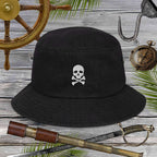 Black bucket hat with skull and crossbones embroidery on a wooden background with pirate-themed items.