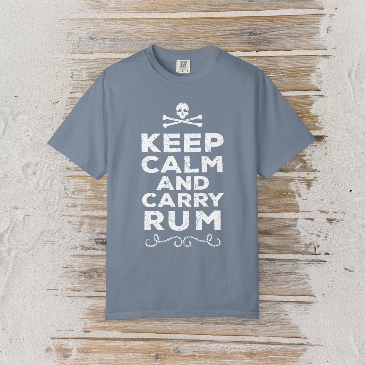 A slate blue crewneck t-shirt with a white graphic print reading 'KEEP CALM AND CARRY RUM' below a skull and crossbones, displayed flat on a rustic wooden plank background.